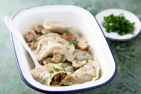 Pyszne pierogi z pieczarkami i serem, posypane natką pietruszki, w białej misce z niebieską obwódką.