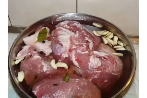Mięso wieprzowe w misce z wodą, czosnkiem, zielem angielskim i liściem laurowym. To przygotowanie do peklowania, czyli solanka do boczku.