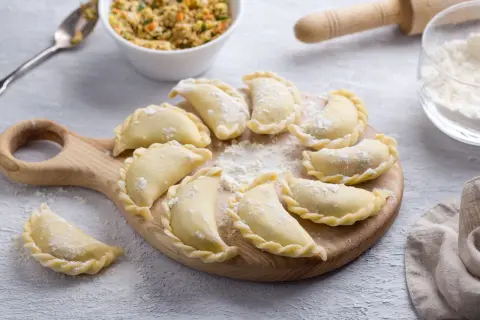 Gotowe do gotowania pierogi ruskie, ułożone na desce. Obok miska z farszem, mąka i wałek. Idealny przepis na pierogi ruskie.