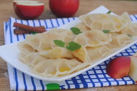 Pyszne pierogi z jabłkami, posypane cynamonem, z listkami mięty i kawałkami jabłek w tle.