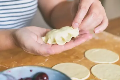 Dłonie formują pieroga z ciasta na pierogi bez jajka. Na blacie leżą okrągłe placki ciasta i miseczka z wiśniami.