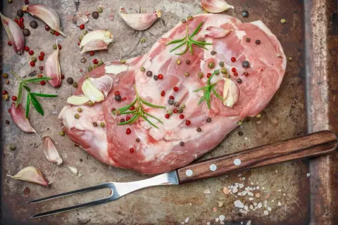 Surowe mięso, czosnek, rozmaryn i kolorowe ziarna pieprzu na metalowej tacy. Widać piękny, różowy kolor mięsa po peklowaniu.