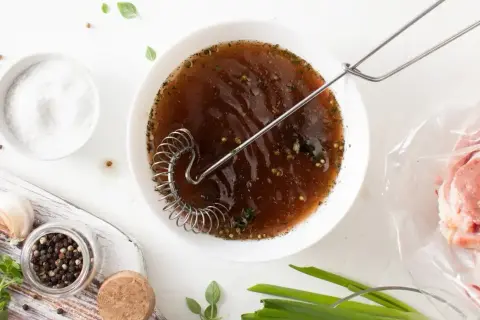 Przygotowanie marynaty do mięsa w solance. W misce brązowy płyn z ziołami i przyprawami, obok sól, pieprz, czosnek i surowe mięso w woreczku.