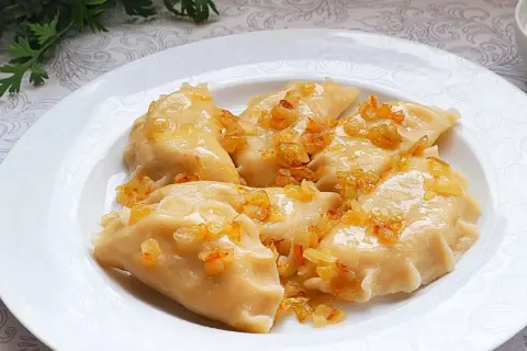 Pyszne pierogi ruskie, polane skwarkami z cebulki, na białym talerzu.