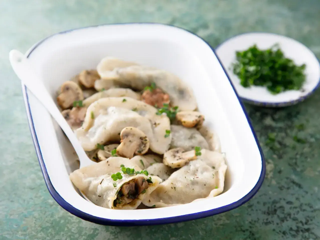 Pyszne pierogi z pieczarkami i serem, posypane natką pietruszki, w białej misce z niebieską obwódką.