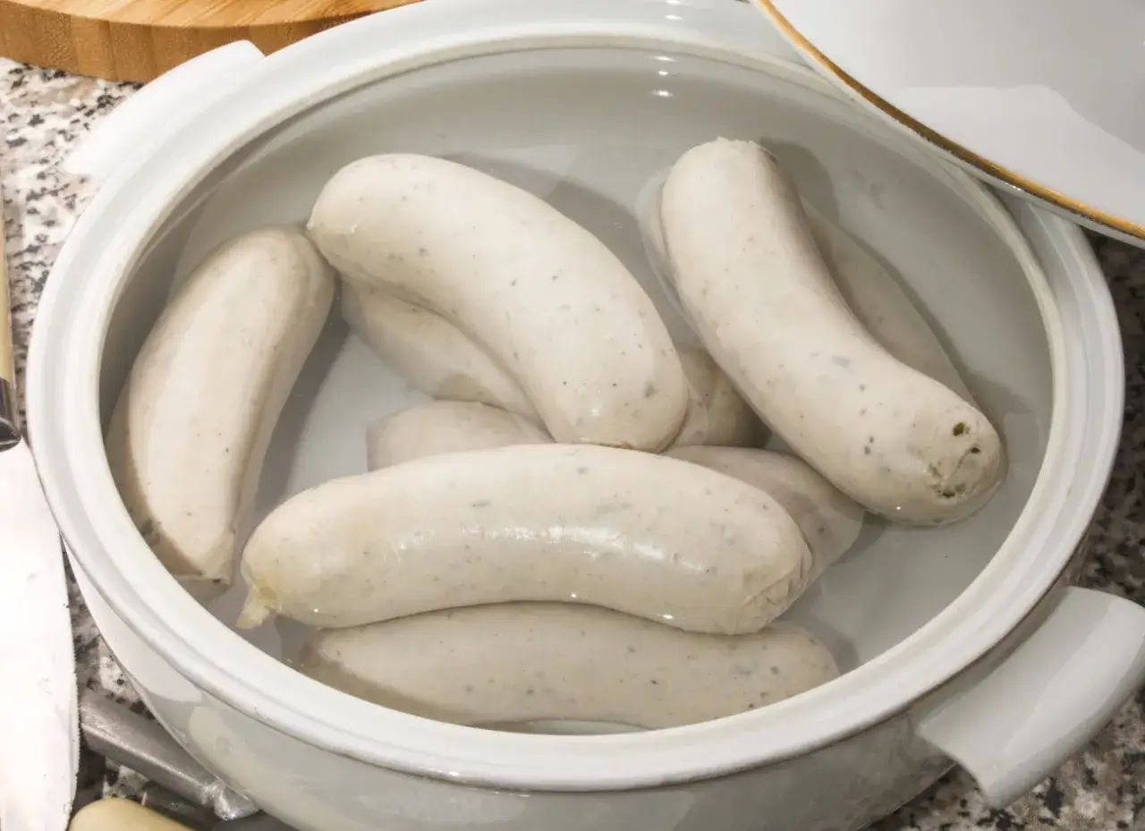 Kilka białych kiełbasek w garnku z wodą, gotowych do parzenia.